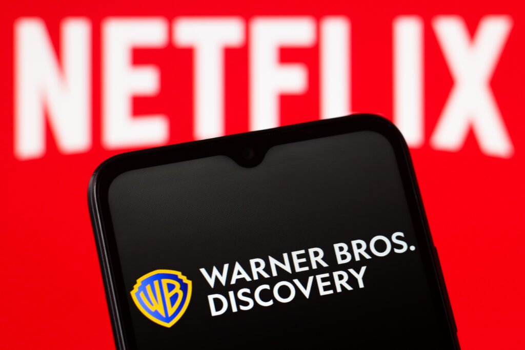 in this photo illustration, the warner bros. discovery logo is seen displayed on a smartphone screen and the logo of netflix in the background.